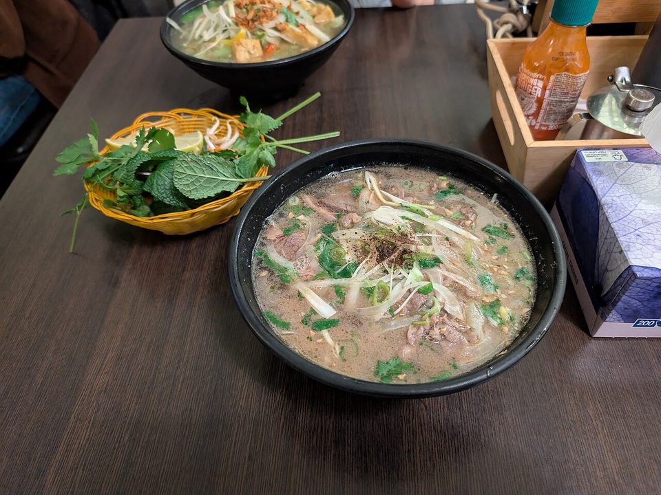 Schale Phở bò mit Rindfleisch, Reisnudeln und Kräutern, daneben ein Kräuterteller